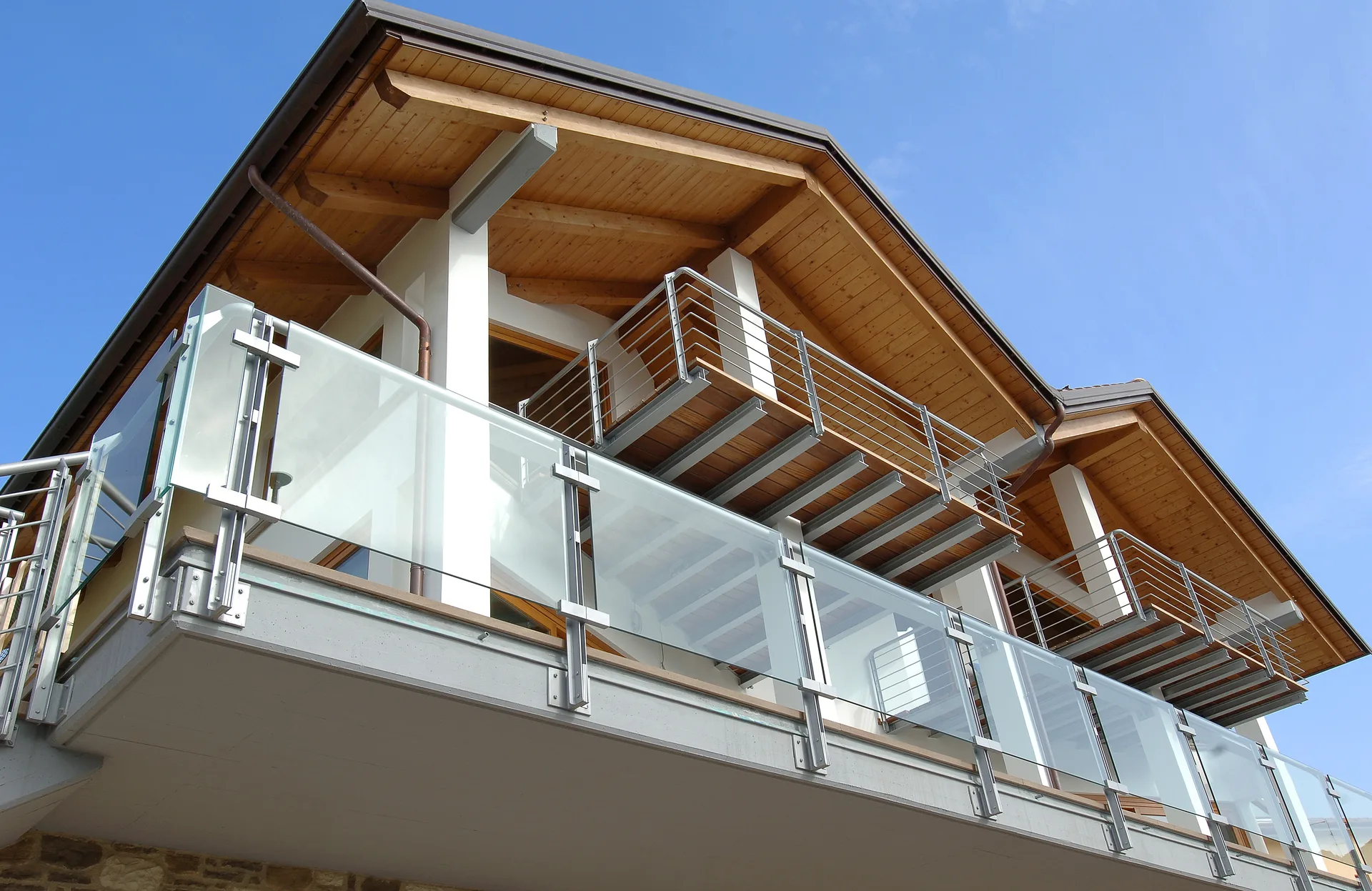 Villa moderna con tetto e coperture in legno lamellare a vista, pilastri strutturali bianchi e ampi terrazzi con ringhiere in vetro e acciaio.